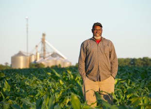 Three-quarter length selective focus view of a mature African-American male farmer standing in a corn field and smiling at the camera